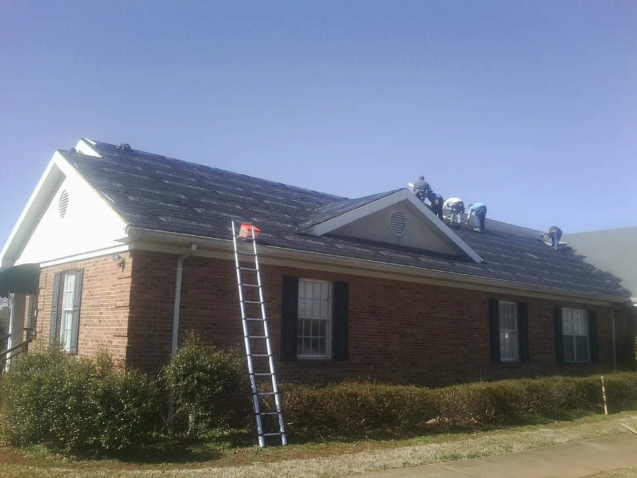 Crew tearing shingles off a brick ranch roof with a ladder set — Emergency Roof Repair work by our crew in Camp Douglas