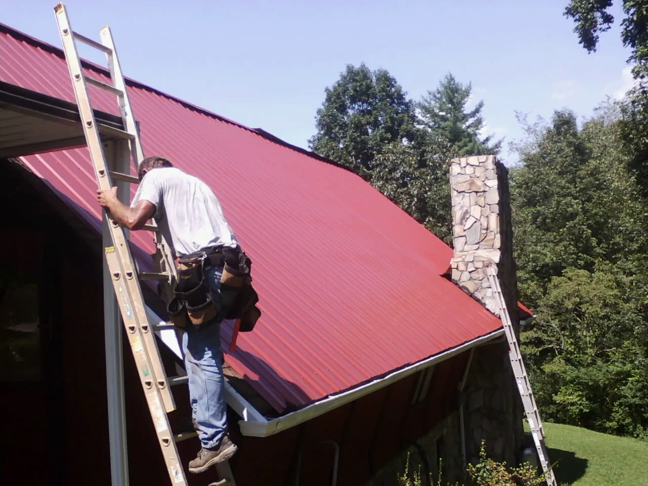 Roofer on a ladder beside a red metal roof and stone chimney — Soffit & Fascia Repair by Roofing Brothers in Camp Douglas